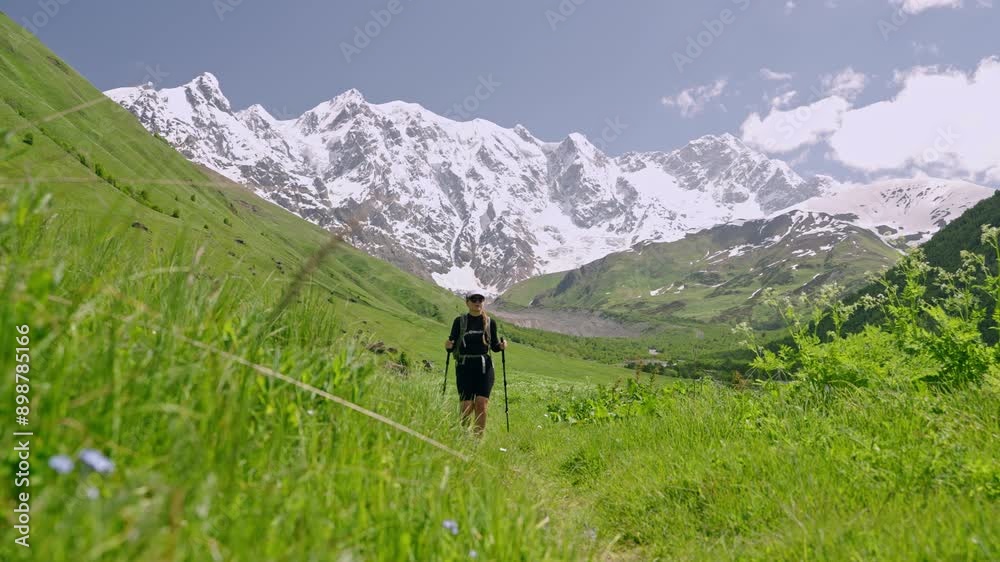 Female tourist hikes with backpack in Jangi-Tau valley, Svaneti. Woman explores nature trail with trekking poles amidst green hills and snowy mountains under clear blue sky. Slow motion. Female tourist hikes with backpack in Jangi-Tau valley, Svaneti. Woman explores nature trail with trekking poles amidst green hills and snowy mountains under clear blue sky. Slow motion.
