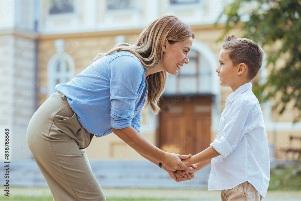 Obraz premium A smiling woman and her young son engage in a loving exchange by a school, reflecting a moment of daily family life with their casual attire and backpacks.
