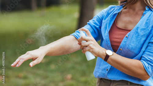 A Caucasian woman dressed casually applies mosquito spray to her arm outdoors, ensuring skin protection in a natural setting.