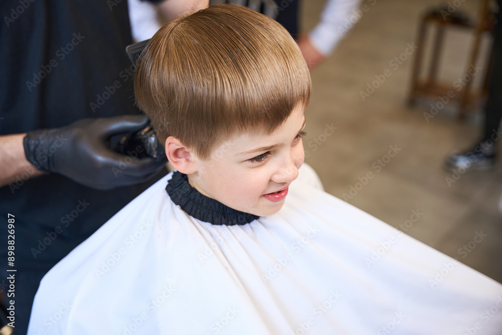 Little client of barbershop waiting hairdresser using trimmer to cut his hair Little client of barbershop waiting hairdresser using trimmer to cut his hair