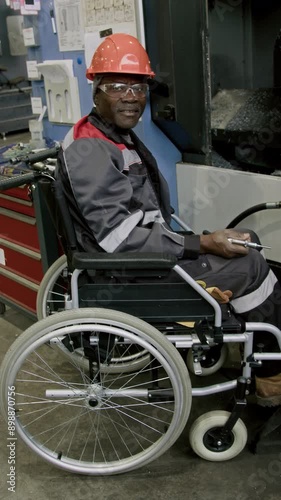 Vertical full portrait of smiling male African American workman in hardhat, overalls and protective goggles posing in factory workshop in wheelchair with air blow gun in hands photo