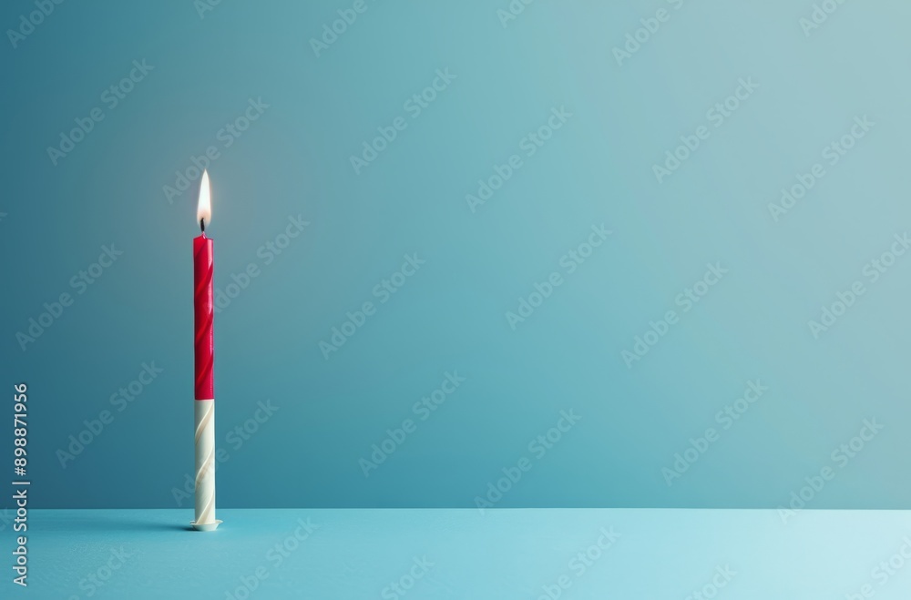A single birthday candle red and white is standing upright on the left side of an empty room, with its flame burning brightly against a plain blue background. A single birthday candle red and white is standing upright on the left side of an empty room, with its flame burning brightly against a plain blue background.