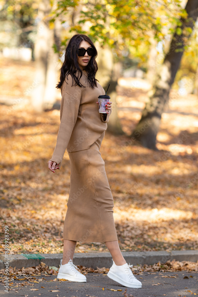 Attractive stylish woman walking in park with a cup of coffee, dressed in warm brown suit and sunglasses. Girl on a background of golden leaves. Autumn fashion accessories in street fashion style Attractive stylish woman walking in park with a cup of coffee, dressed in warm brown suit and sunglasses. Girl on a background of golden leaves. Autumn fashion accessories in street fashion style