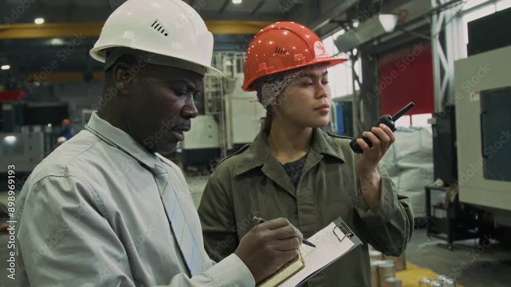Side medium slowmo of mature male African American supervisor in shirt and tie making notes in report while Latin forewoman giving orders via walkie-talkie, controlling quality of production Side medium slowmo of mature male African American supervisor in shirt and tie making notes in report while Latin forewoman giving orders via walkie-talkie, controlling quality of production