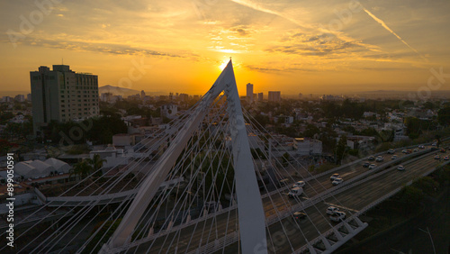 A majestic sunrise over the Matute Remus Bridge in Guadalajara, Mexico, casts a warm orange glow across the city, illuminating the sleek structure and creating a stunning visual contrast against the a photo