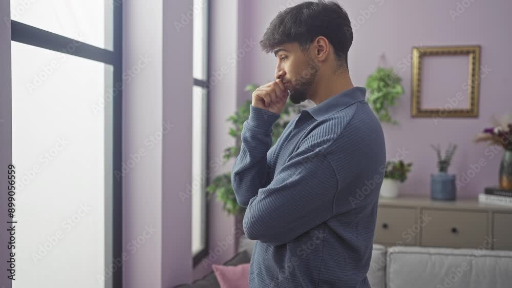 A contemplative young bearded man standing in a modern apartment living room, looking thoughtful and serene.