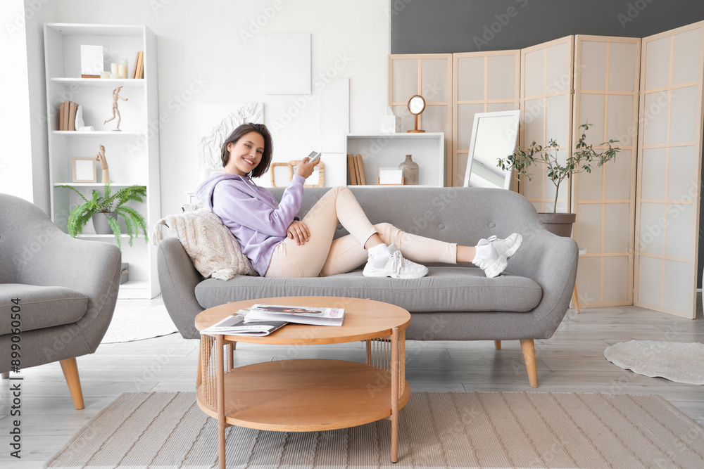 Smiling young woman with air conditioner remote control lying on sofa in living room Smiling young woman with air conditioner remote control lying on sofa in living room