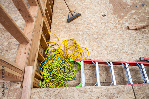 View looking down at basement subfloor, Oriented Strand Board, house new construction wood framing with ladder, push broom, and pile of air compressor hoses stored on floor
