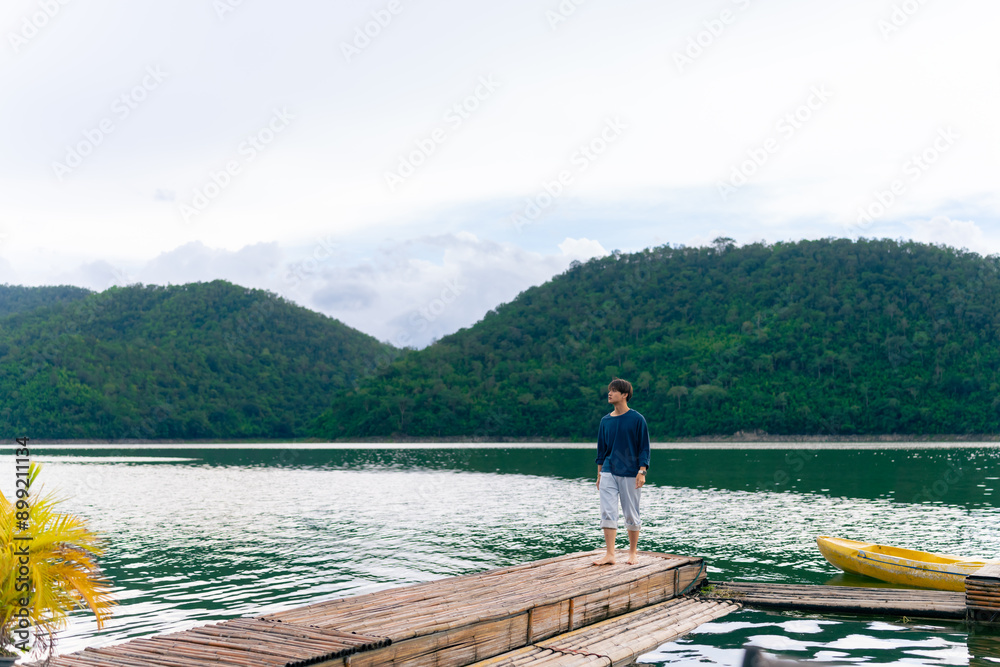Fototapeta premium Young Asian man relax and enjoy outdoor lifestyle travel forest mountain on summer holiday vacation. Generation z guy relaxing on lake house balcony looking beautiful nature and breathing fresh air