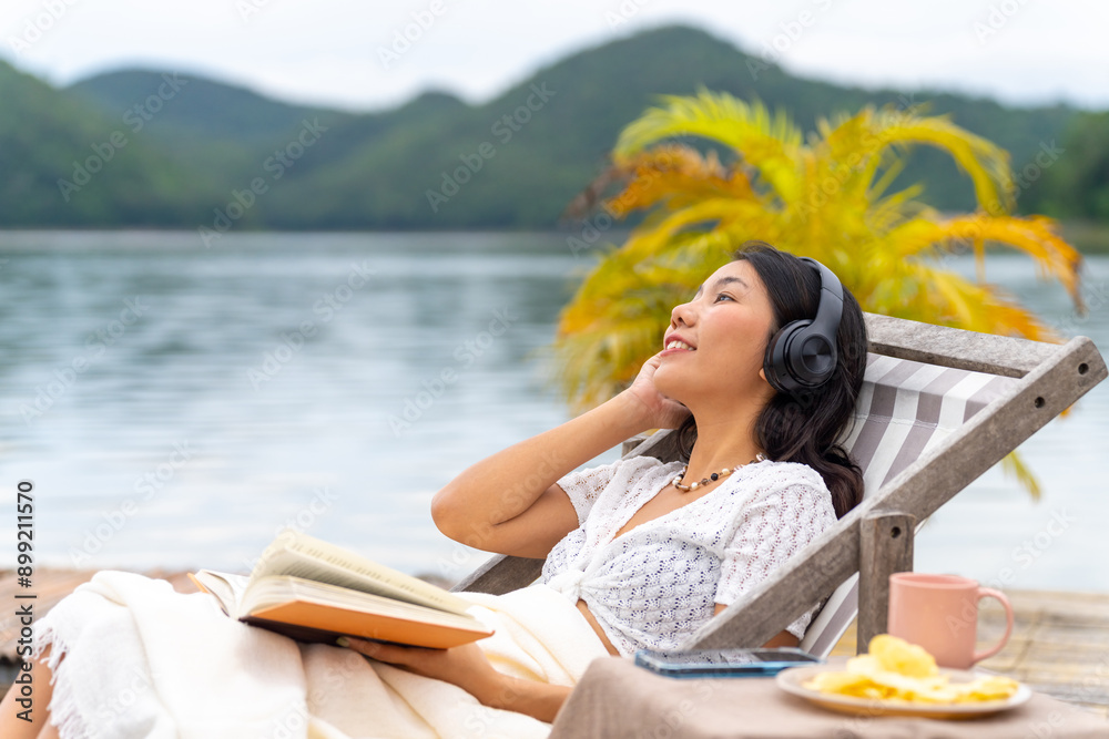 Fototapeta premium Asian woman sitting on lake house balcony reading a book and listening to the music on headphones with mobile app. Attractive girl enjoy outdoor lifestyle travel nature on summer holiday vacation.