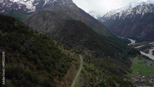 Winding road through steepy mountain range covered with dense forest, snowy mountain peaks in background and river flowing below near small village. Aerial view photo