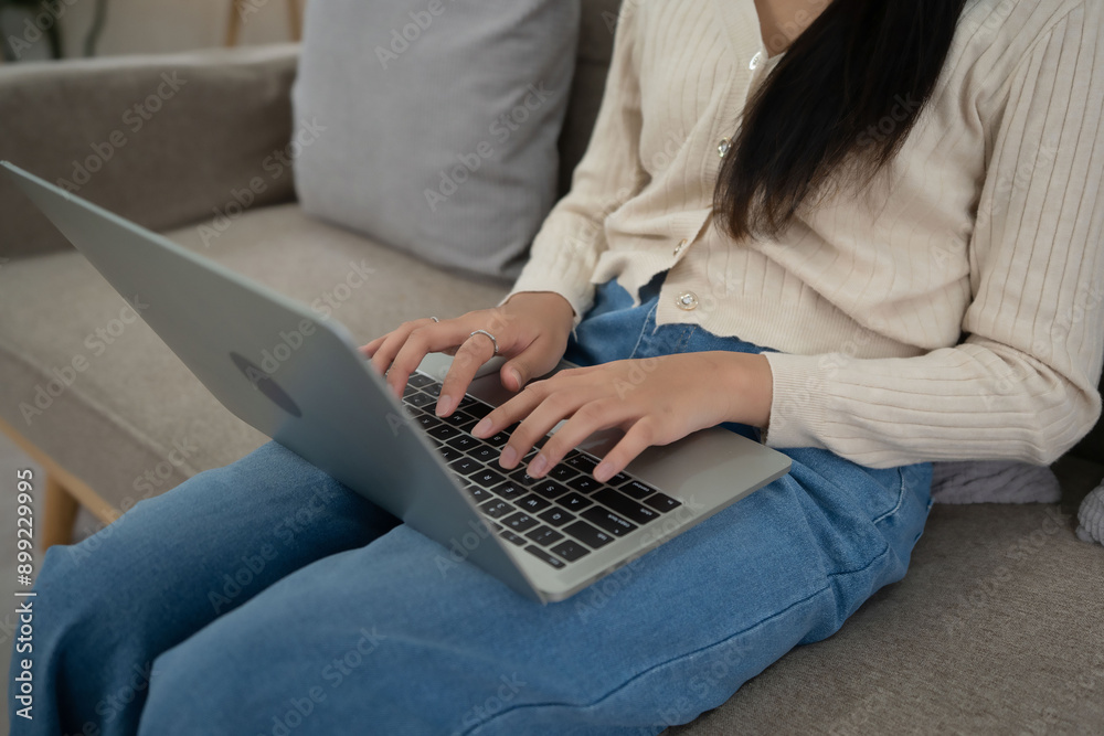 Fototapeta premium Closeup of young asian woman putting laptop on legs and typing business report about marketing data of project while sitting on couch to spending time and relaxation with enjoying lifestyle at home