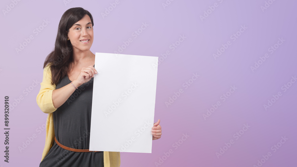Happy mid adult female entrepreneur dressed in grey dress and yellow jacket showing white blank placard template mock up while standing isolated on purple background Happy mid adult female entrepreneur dressed in grey dress and yellow jacket showing white blank placard template mock up while standing isolated on purple background