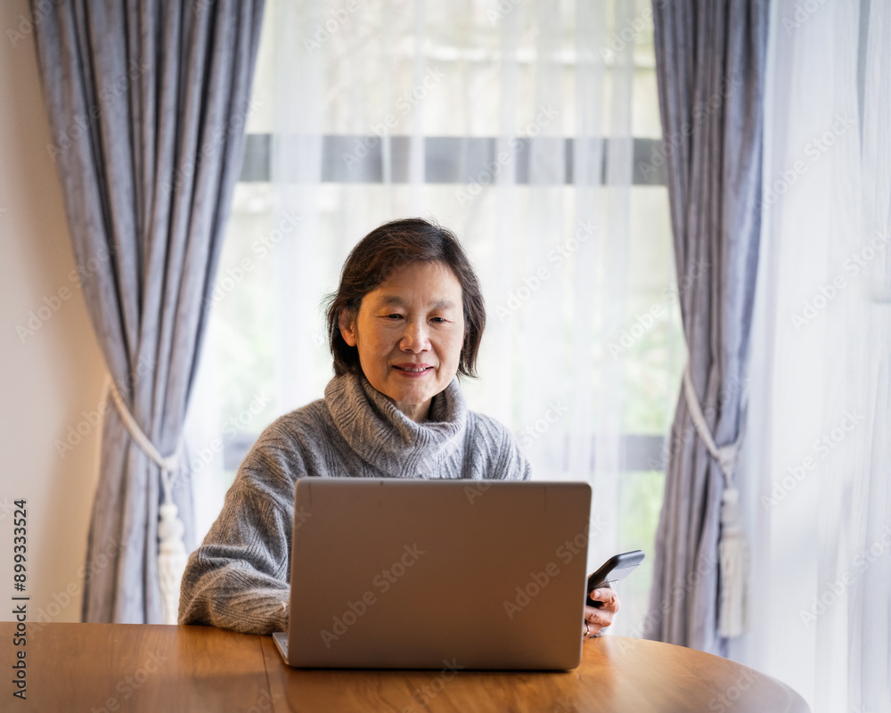Senior woman smiling and reading from laptop at home, left hand holding a smartphone.