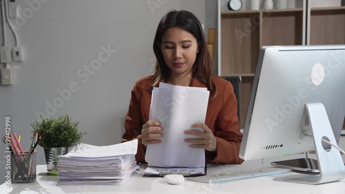 An Asian businesswoman sits at a table, working with a smile. She manages her online Kuya business with enthusiasm, showcasing her dedication and positive attitude while handling tasks efficiently. photo