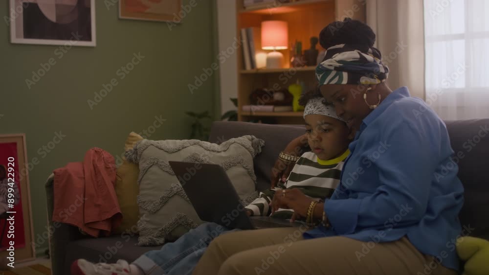 Medium long shot of child and mother of African American ethnicity watching videos on laptop when sitting on sofa at evening Medium long shot of child and mother of African American ethnicity watching videos on laptop when sitting on sofa at evening