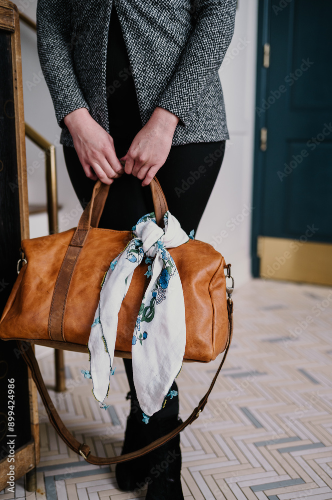 Fototapeta premium Woman holding a brown leather bag with a floral scarf tied to it.