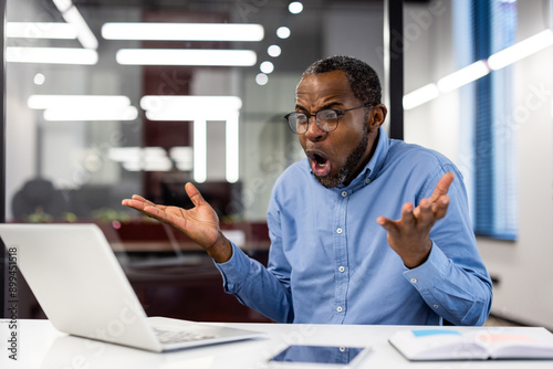 Surprised businessman sitting at desk with laptop, experiencing shock or disbelief. Bright modern office setting, professional attire. Concept of unexpected news, business challenge