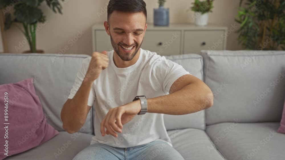 A young hispanic man in a living room smiles while looking at his smartwatch, wearing a white t-shirt and sitting on a grey sofa with plants and decor in the background. A young hispanic man in a living room smiles while looking at his smartwatch, wearing a white t-shirt and sitting on a grey sofa with plants and decor in the background.
