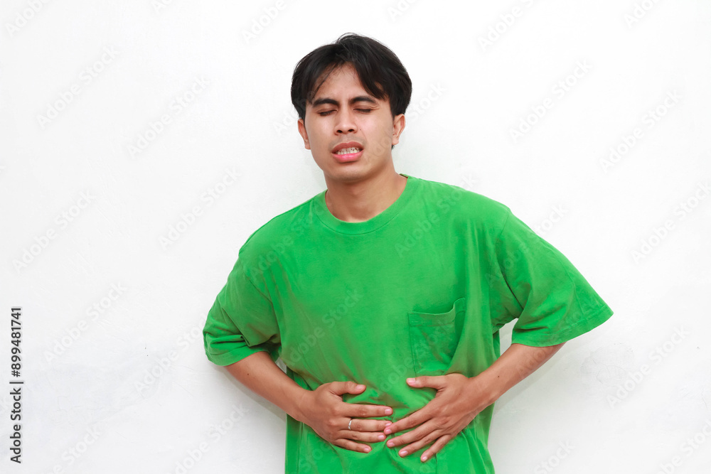 Obraz premium Adult Asian man wearing green shirt posing with gesture of holding body part and showing pain expression on white background.