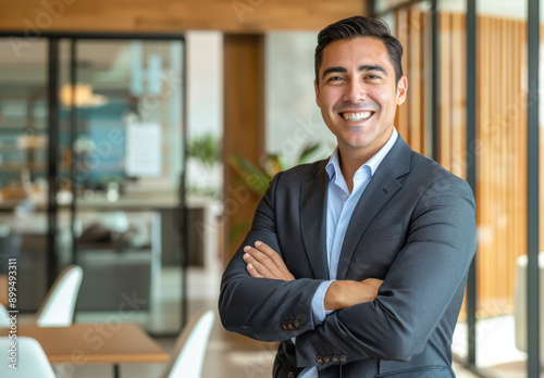 Confident Business Leader in Office Setting. A young Hispanic male, light skin, with black hair, dressed in a professional grey suit, smiling confidently in a corporate environment  © PixelGallery