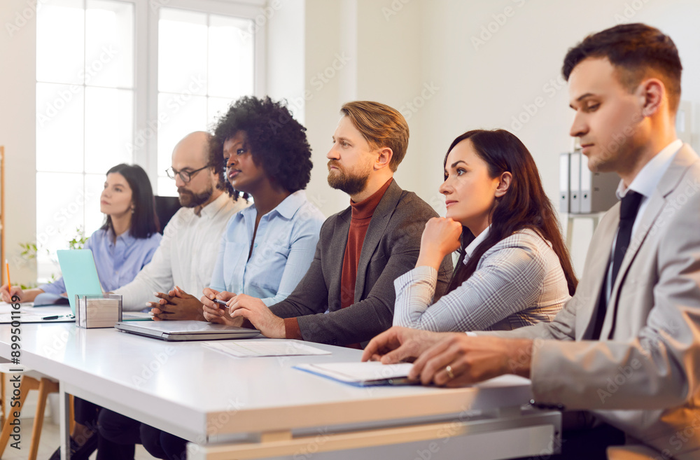 Group of confident diverse business people sitting in a row at desk in meeting room and listening speaker on a conference. Successful coworkers and company employees on business training Group of confident diverse business people sitting in a row at desk in meeting room and listening speaker on a conference. Successful coworkers and company employees on business training