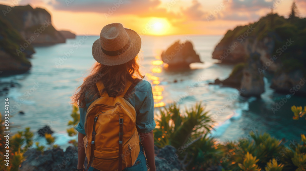 Fototapeta premium Back view of a traveler with a backpack and hat admiring a beautiful sunset over the ocean, surrounded by lush greenery.