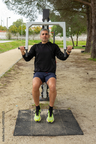 A gray-haired man works out on a chest press machine at an outdoor fitness station in an urban park. He exercises with determination, enjoying the fresh air and staying active. photo