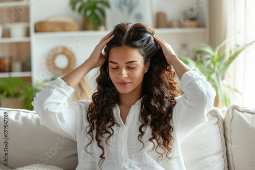 Young woman is sitting on a comfortable sofa, gently massaging her scalp with her eyes closed, enjoying a moment of relaxation photo