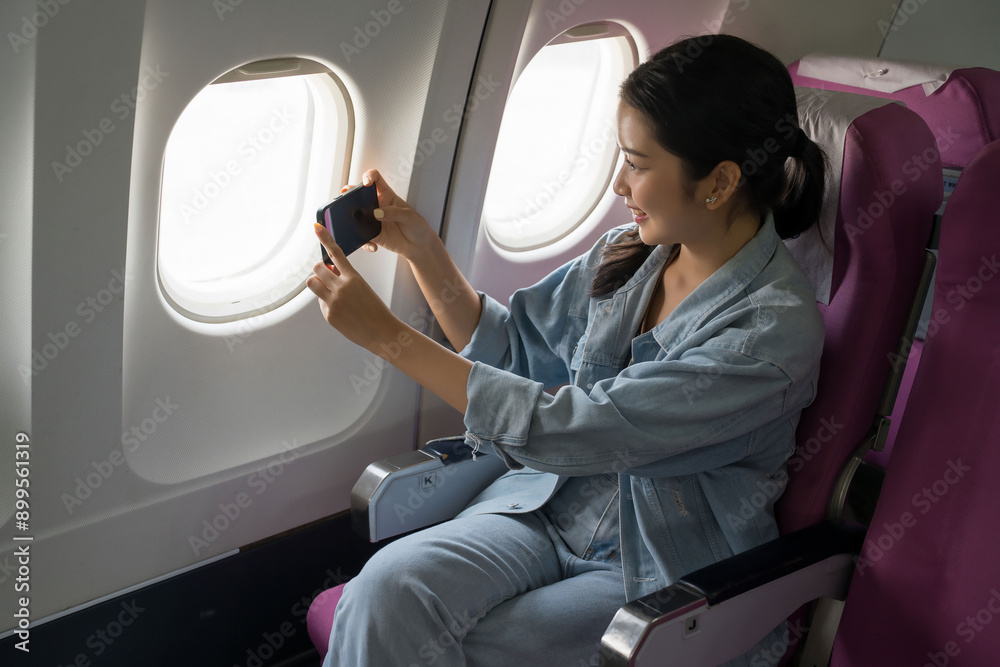 Fototapeta premium Asian traveler women looking and taking photo of sky view on smartphone through the windows on cabins flight with happiness while sitting near windows on business travel class flight in airplane
