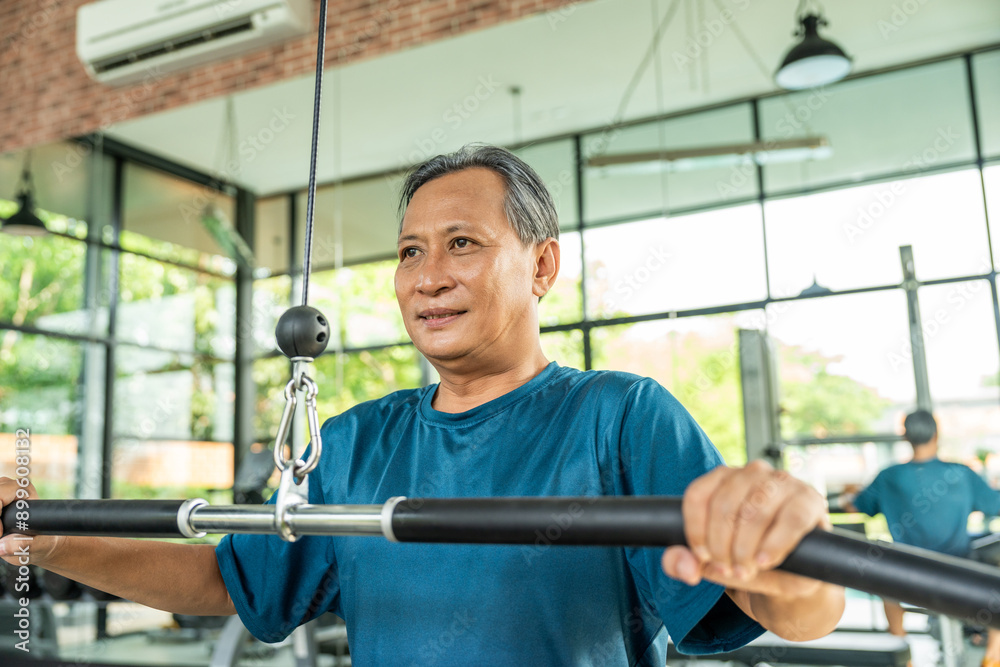 Fototapeta premium Old man weight training wearing sportswear. Asian mature exercise indoor gym. Bodybuilder good health. Fitness elderly pull up the machine bar in gym.