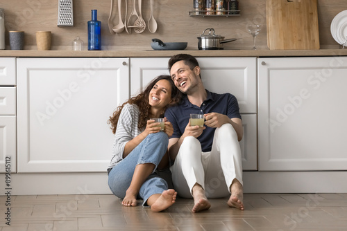 Young spouses drinking juice relax together seated on warm laminate floor with underfloor heat system in kitchen, look away, daydream, share plans and dreams on relocation day to their new modern flat