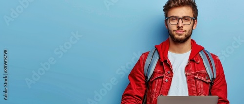 Young man with glasses holding a laptop against a blue background, wearing a red jacket and backpack, looking at the camera.