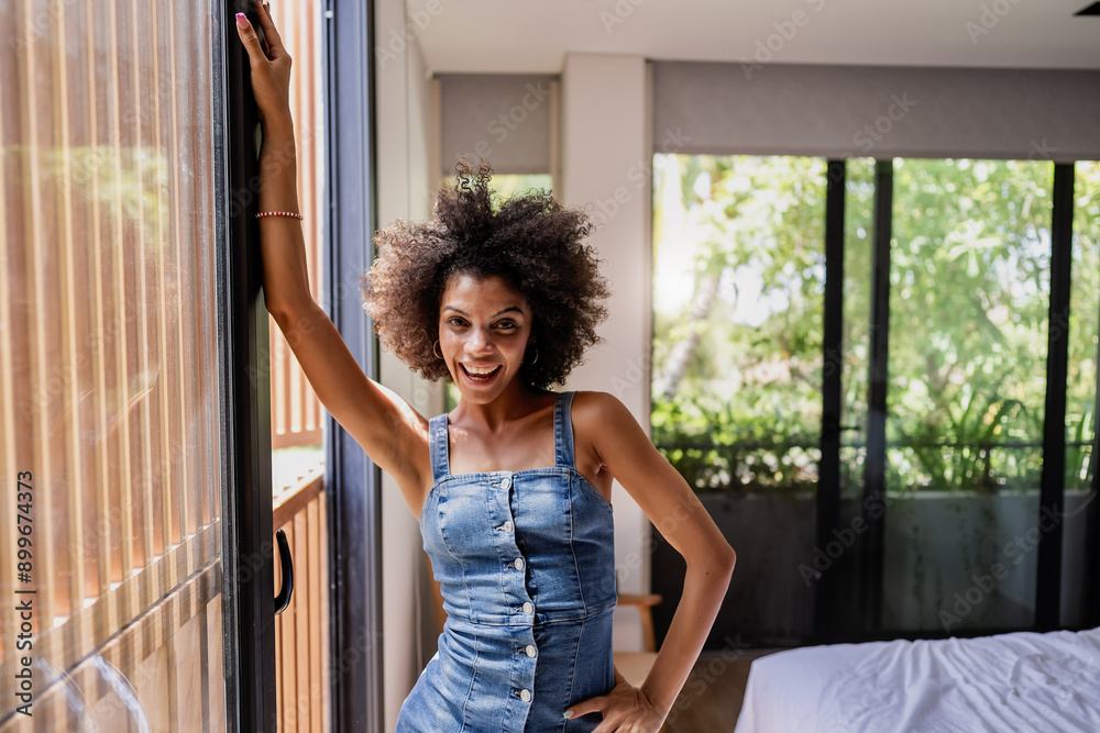 Fototapeta premium Portrait of a young beautiful cheerful woman of African descent standing near a big sunlit window of a hotel room.
