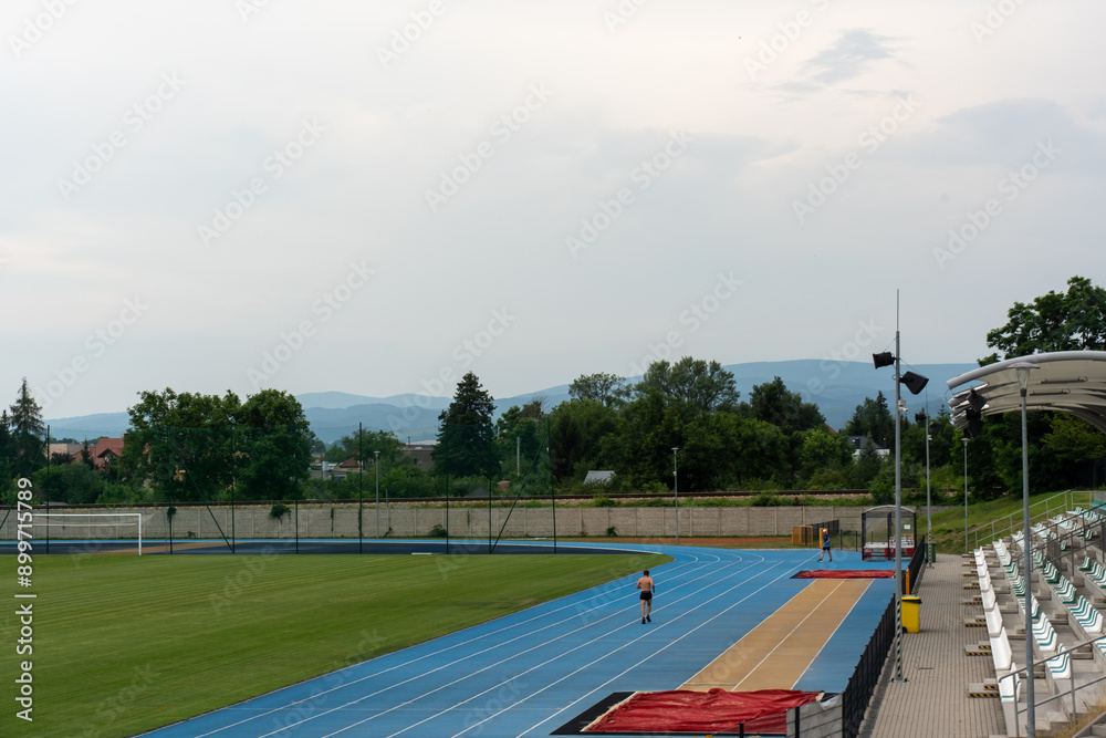 Fototapeta premium Lone runner on blue track at outdoor stadium with lush green field and scenic mountain backdrop on cloudy day. Concept of athletic training, outdoor exercise, and sports venues. High quality photo