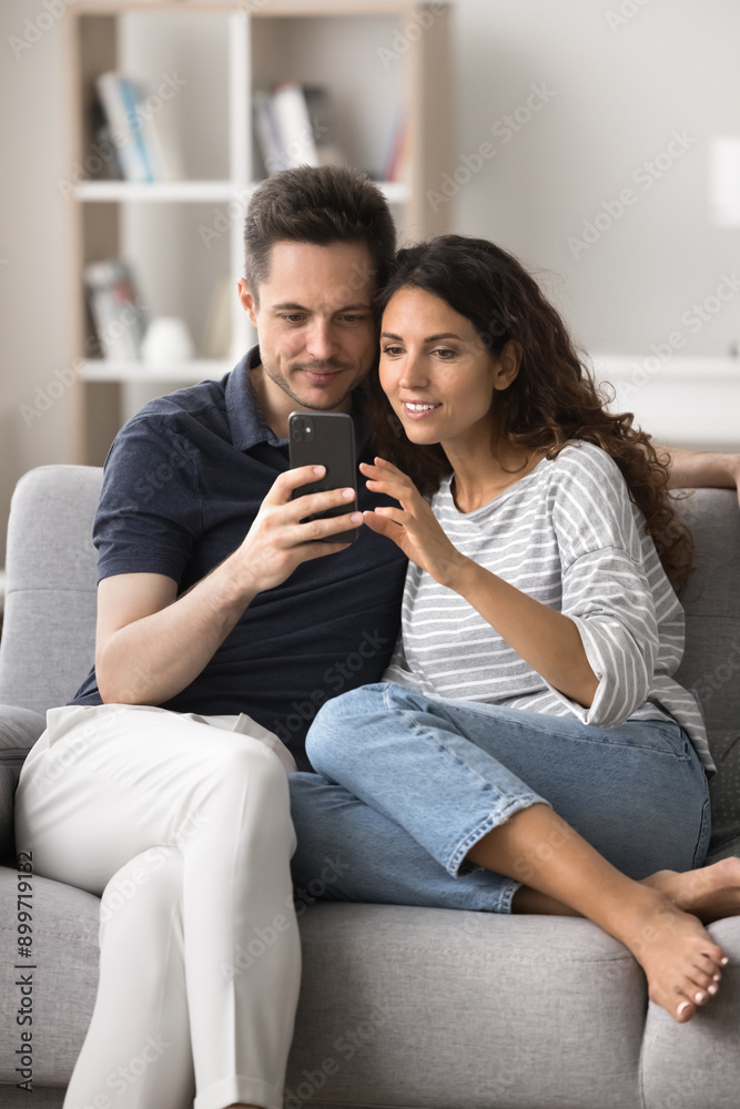 Fototapeta premium Hispanic spouses rest on cozy sofa looking at mobile phone screen, web surfing information, buy tickets, booking hotel, enjoy new modern application, communicate using wireless internet. Technology