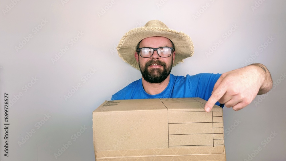 Portrait of bearded handsome delivery man in straw hat and blue shirt delivering box package, isolated on white background