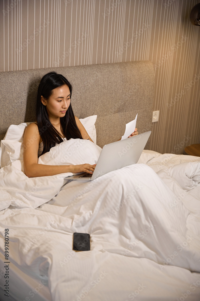Focused Asian woman sitting in bed and working on diploma project Focused Asian woman sitting in bed and working on diploma project
