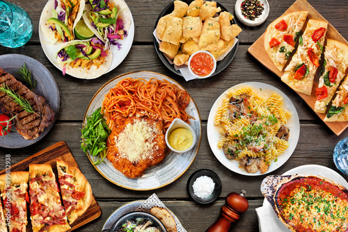 Table scene of various delicious foods. Top down view on a dark wood background. Pastas, flatbreads, tacos, steak and restaurant theme dishes.