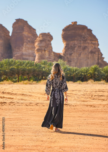 Young woman wearing long dark dress, walking on desert landscape, palm trees and large rock formation typical for Al Ula region in distance. View from behind