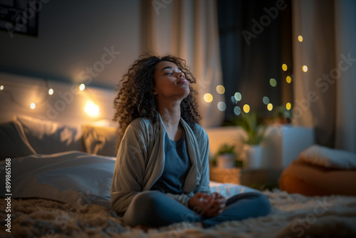 A woman in cozy sleepwear sits cross-legged on her bed, meditating amidst a softly lit bedroom. String lights and indoor plants create a peaceful, serene atmosphere,relaxation and mindfulness. photo