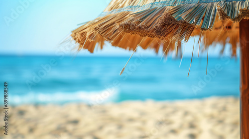 A coconut straw umbrella set on the beach where the sand meets the ocean, with a blurry blue sky in the background. The exotic view of a tropical island creates the perfect setting for a summer vacati photo