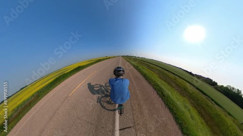 Action camera following a man in a blue shirt that is pedaling a bicycle along the centre line of a paved road that runs through farm fields.
 photo