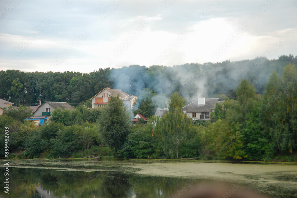 Fototapeta premium Smoke over private houses over the lake and green trees.