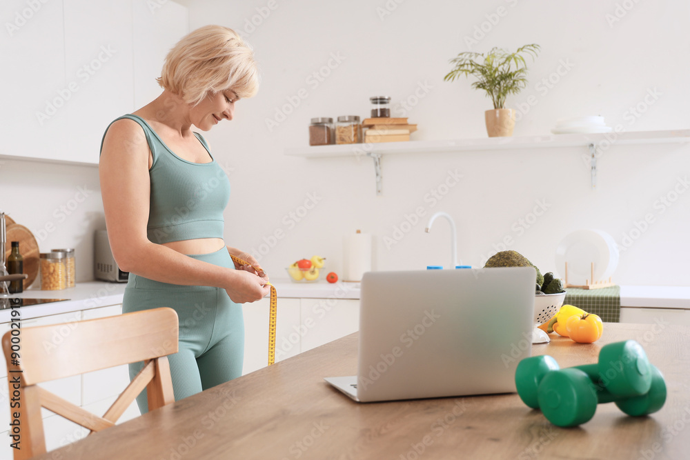 Middle-aged woman with measuring tape, dumbbells and laptop in kitchen Middle-aged woman with measuring tape, dumbbells and laptop in kitchen