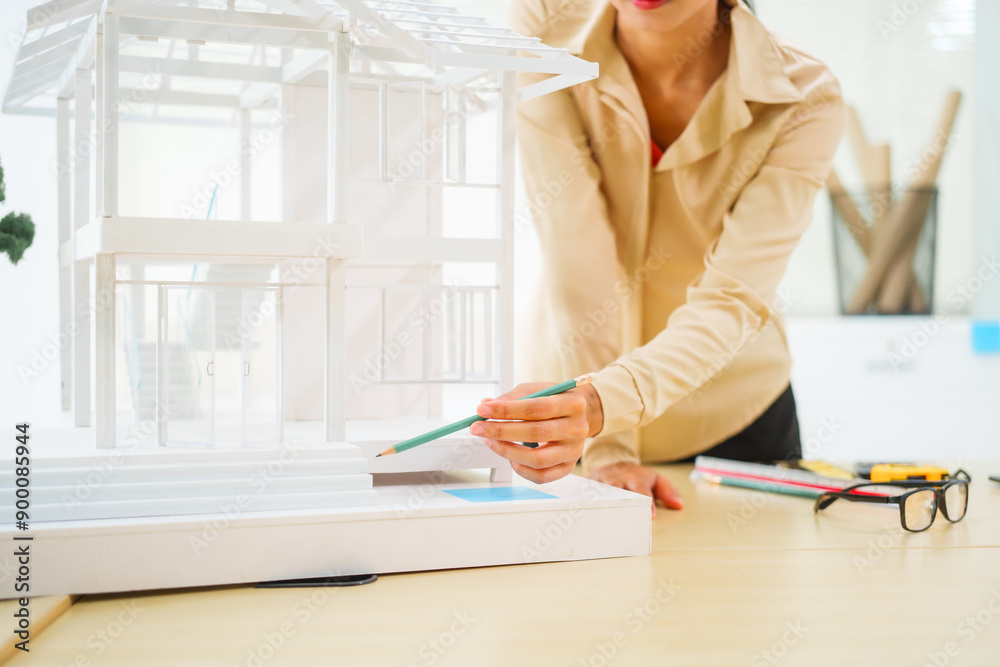 An Asian female architect analyzes blueprints at her desk in the office. She specializes in house design, focusing on roofs and walls, and collaborates with construction contractors in her company. An Asian female architect analyzes blueprints at her desk in the office. She specializes in house design, focusing on roofs and walls, and collaborates with construction contractors in her company.