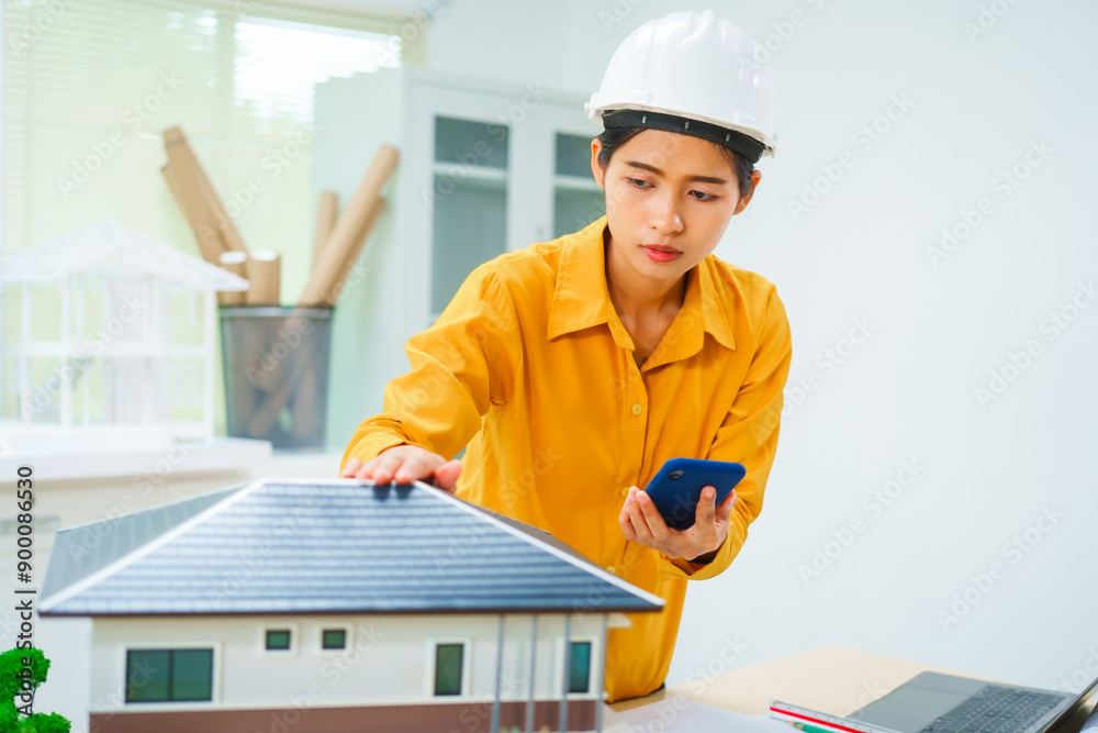 An Asian female architect analyzes blueprints at her desk in the office. She specializes in house design, focusing on roofs and walls, and collaborates with construction contractors in her company.