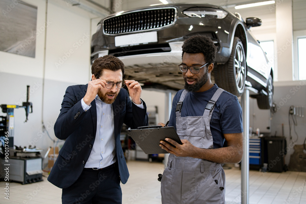 Businessman in suit consulting with mechanic in auto workshop. Mechanic in overalls holding clipboard explaining car repair details. Modern garage with vehicle lifted in background. Businessman in suit consulting with mechanic in auto workshop. Mechanic in overalls holding clipboard explaining car repair details. Modern garage with vehicle lifted in background.
