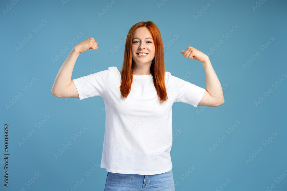 Fototapeta premium Redhead woman showing her strength flexing biceps muscles on blue background