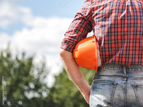 Attractive man in work clothes with a protective helmet against a background of trees and blue sky. Rear view, close-up, outdoors. Labor and employment concept